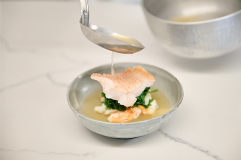 Broth being ladled into a bowl with fish and greens.