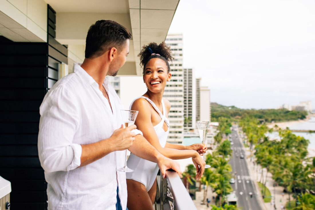 Man and woman enjoying champagne on balcony with views of Kalalakua Avenue and Waikiki beach.